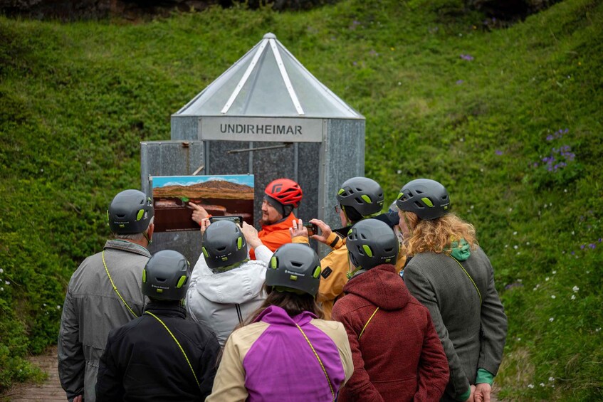 A group of people wearing helmets and jackets gathered around a structure labeled 'UNDIRHEIMAR' with a scenic landscape painting on it. 2