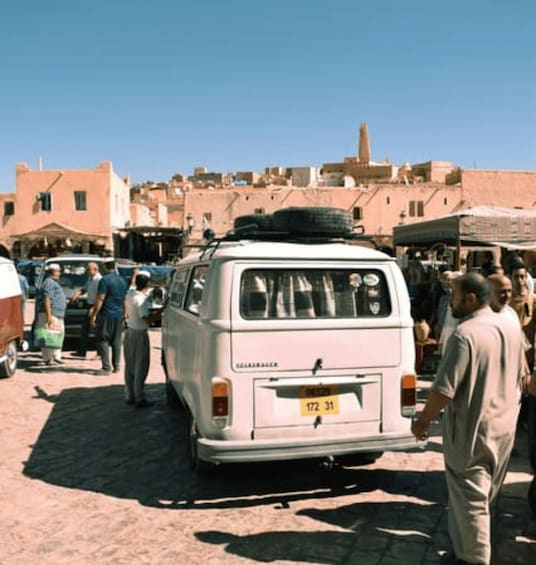 A white van parked on a cobblestone street in a market area with people around. 2