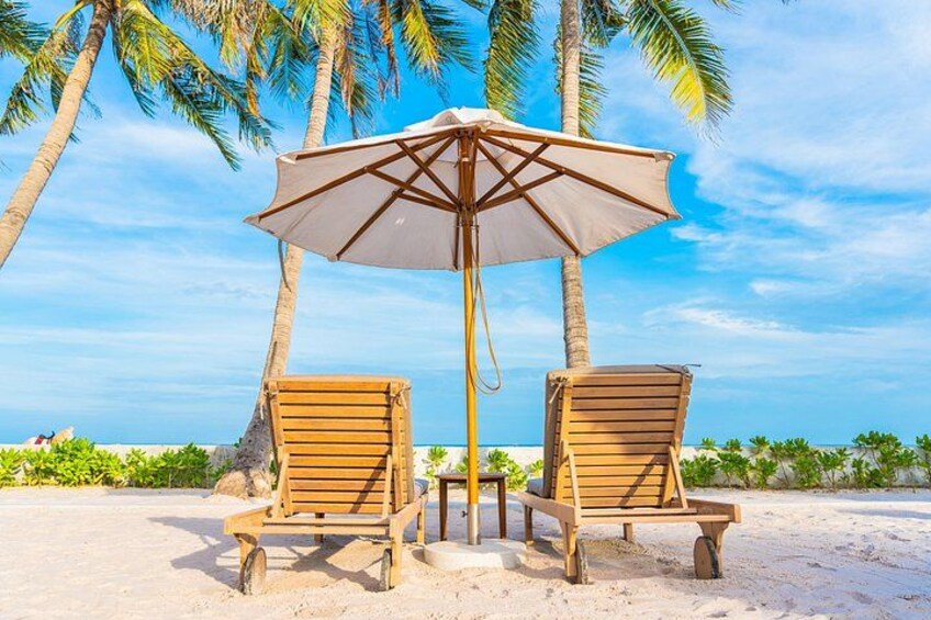 Two wooden beach chairs under a large umbrella on a sandy beach with palm trees in the background. 2