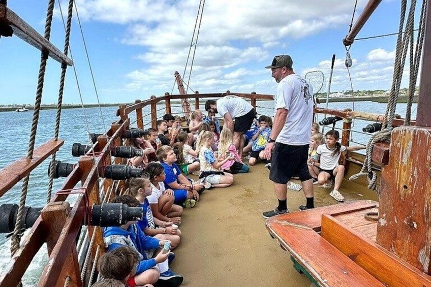Eine Gruppe von Menschen auf einem Boot, einige sitzen, andere stehen, während ein Mann sie anspricht. 4