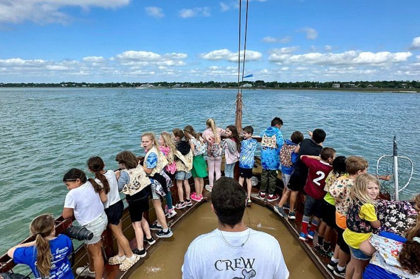 Eine Gruppe von Menschen in einem Boot, einige tragen Rettungswesten, im Hintergrund ruhiges Wasser und ein klarer Himmel. 5