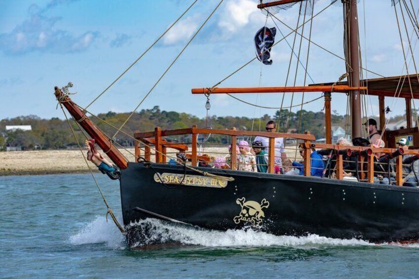 Un barco negro con un logotipo de calavera y tibias cruzadas, llamado 'SEA FURY', navegando sobre agua con una playa arenosa y árboles al fondo. 1