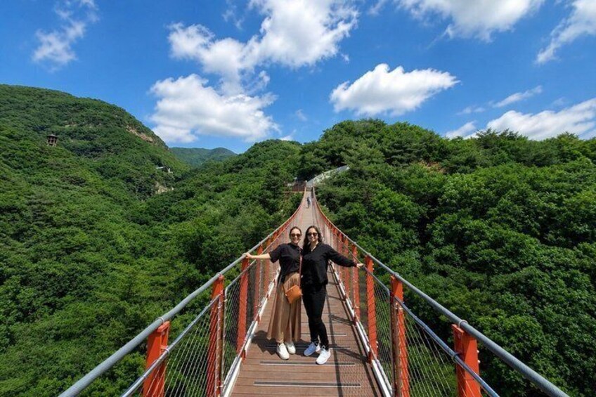 Eine hölzerne Hängebrücke mit Metallgeländern, umgeben von üppigen, grünen Hügeln. 1