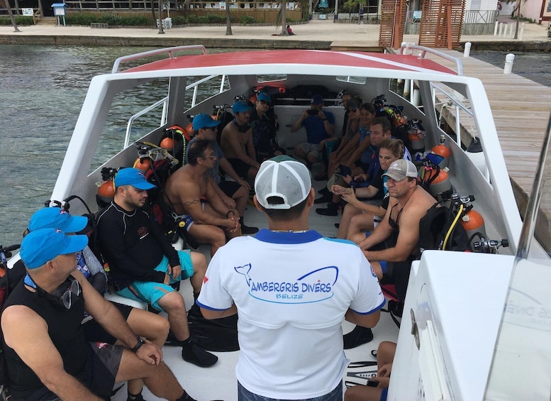 A group of divers on a boat, some wearing diving gear, with a person in a white shirt with 'VAMBERGIRIS DIVERS BELIZE' on the back. 3