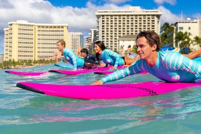 Personas sobre tablas de surf en el agua, palmeras y edificios al fondo. 3