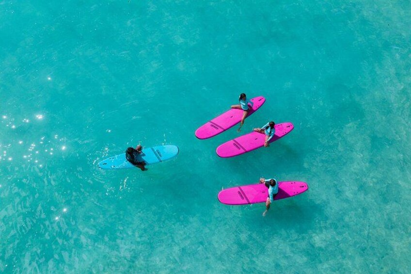 En person på en blå surfbräda och tre personer på rosa surfbrädor i klart, turkosfärgat vatten. 1