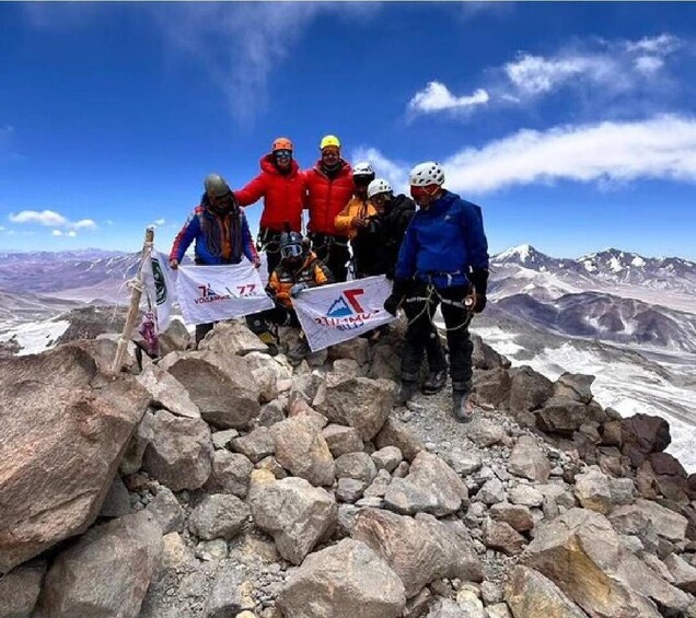 A group of climbers on a rocky mountain peak with flags, under a clear sky. 2