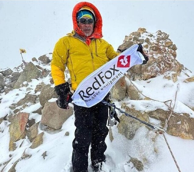A person in a yellow jacket and red hood holding a RedFox outdoor equipment banner on a snowy mountain. 3