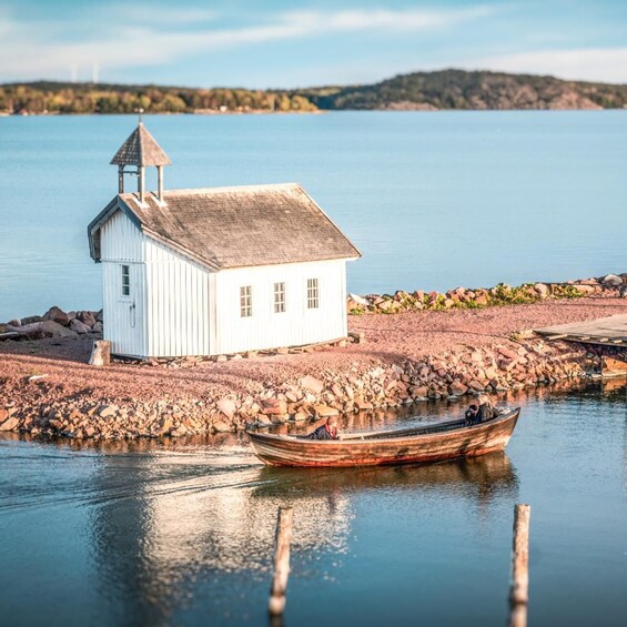 A white wooden structure with a pointed roof on a rocky shore by calm water. 1