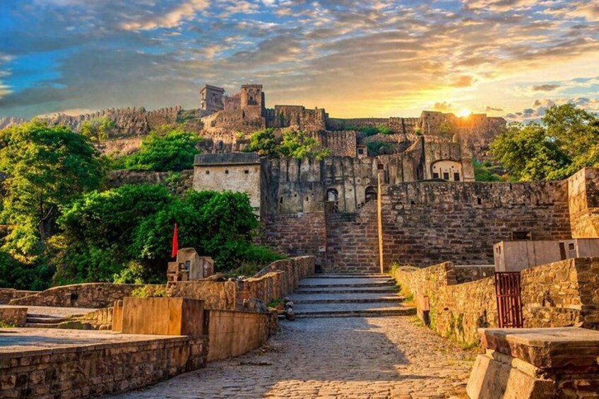 A stone pathway leading to a historic fortress with a flag on the left, surrounded by greenery and a sunset in the background. 4