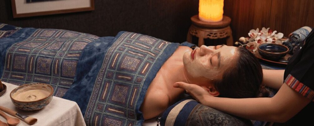 A person lying on a patterned bed with a bowl of cream and a wooden spoon, a candle, and a decorative bowl on a table beside them. 1