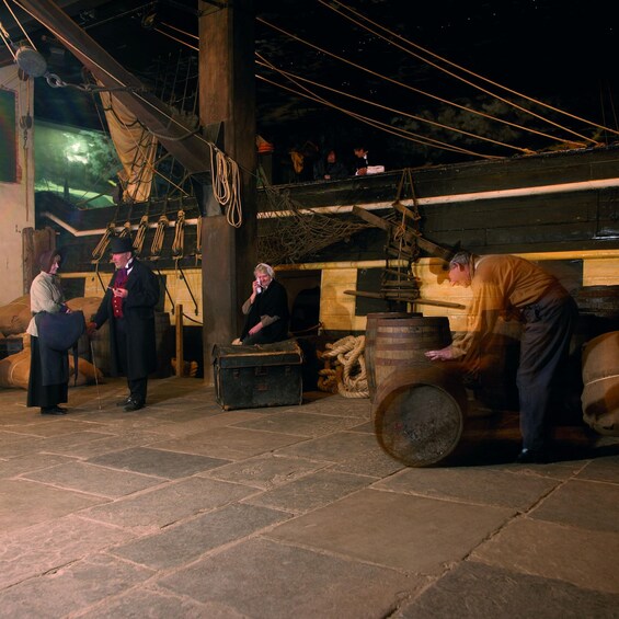 A historical reenactment scene with individuals in period attire, a large wooden barrel, and ropes. 2