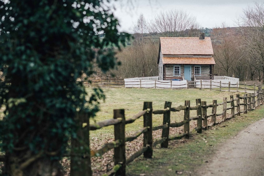 A rustic wooden house with a tiled roof, surrounded by a white picket fence, set in a grassy area with trees in the background. 4