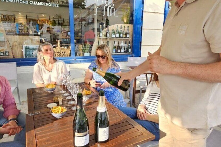 Eine Gruppe von Menschen sitzt an einem Tisch im Freien, mit Champagnerflaschen, einer Obstschale und einem Weinglas. 2