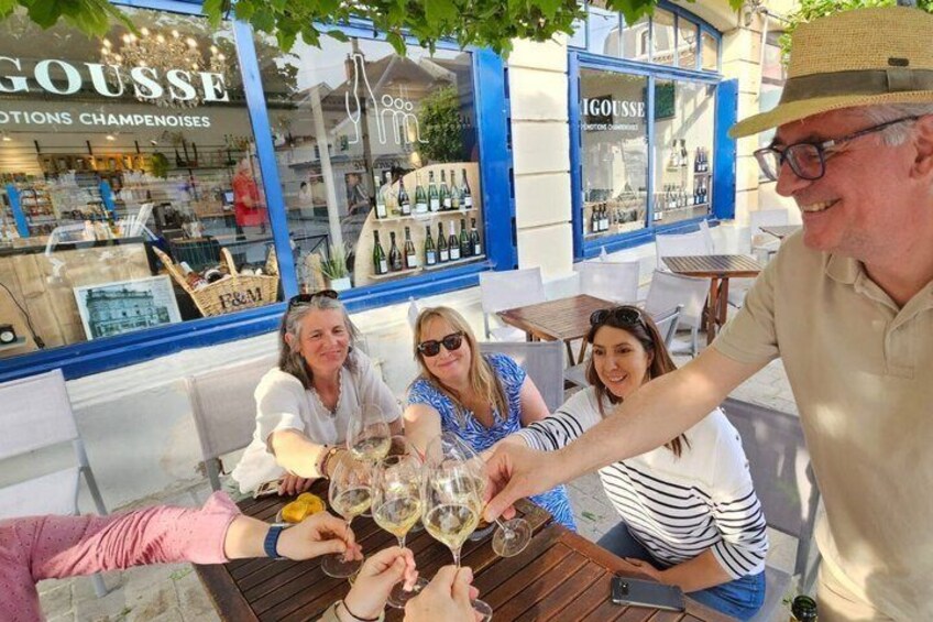 Eine Gruppe von Personen stöstet mit Champagnergläsern in einem Straßencafé mit Blick auf ein Geschäft für Champagner an. 3