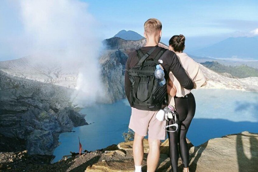 Zwei Personen stehen auf einer felsigen Aussichtsplattform mit einem geothermalen See und Bergen im Hintergrund. 1