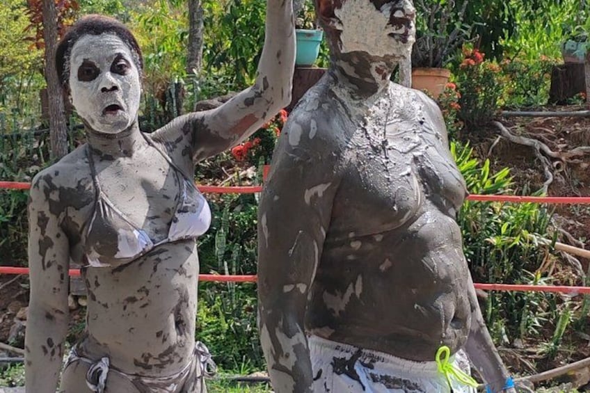 Two statues covered in mud, standing outdoors with greenery and a red fence in the background. 3