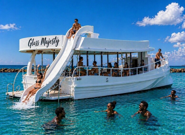 A glass boat named 'Glass Majesty' with a slide, surrounded by clear blue water and people swimming. 1