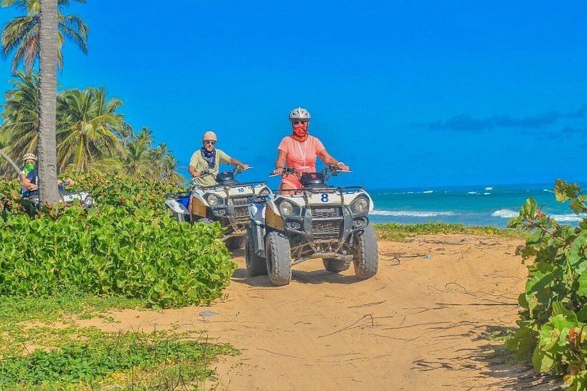 Zwei Personen fahren mit Quads auf einem Sandweg, im Hintergrund sind Palmen und das Meer zu sehen. 2