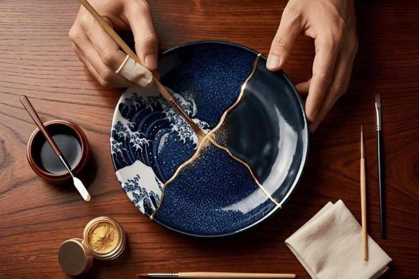 Une personne tient une assiette en céramique bleue cassée, ornée de motifs dorés, ainsi qu’une cuillère en bois, des baguettes et un petit bol de sauce soja, le tout posé sur une table en bois. 4