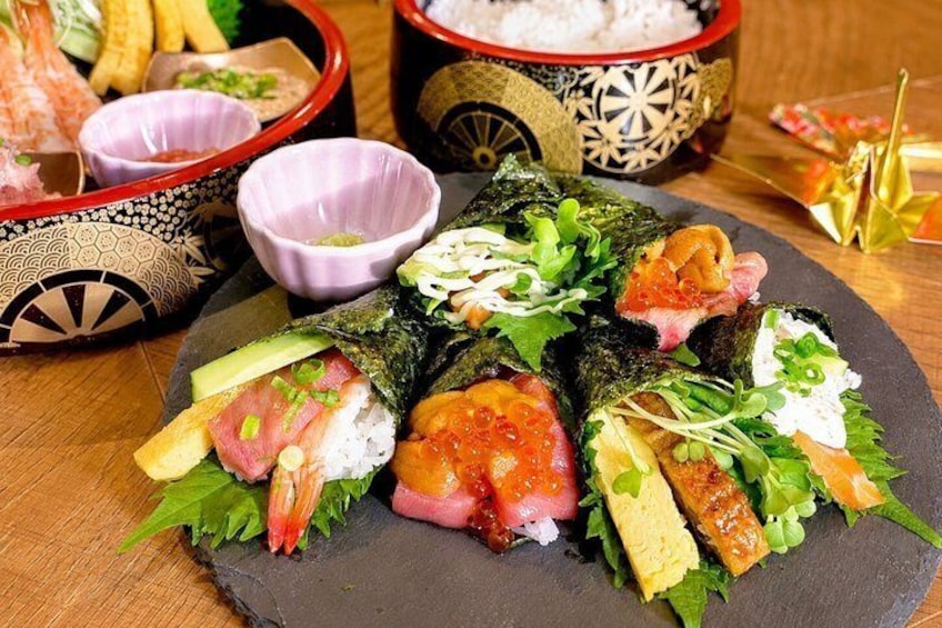 Variedad de rollos de sushi sobre un plato de pizarra, acompañados de recipientes pequeños con condimentos y un cuenco decorativo al fondo. 5
