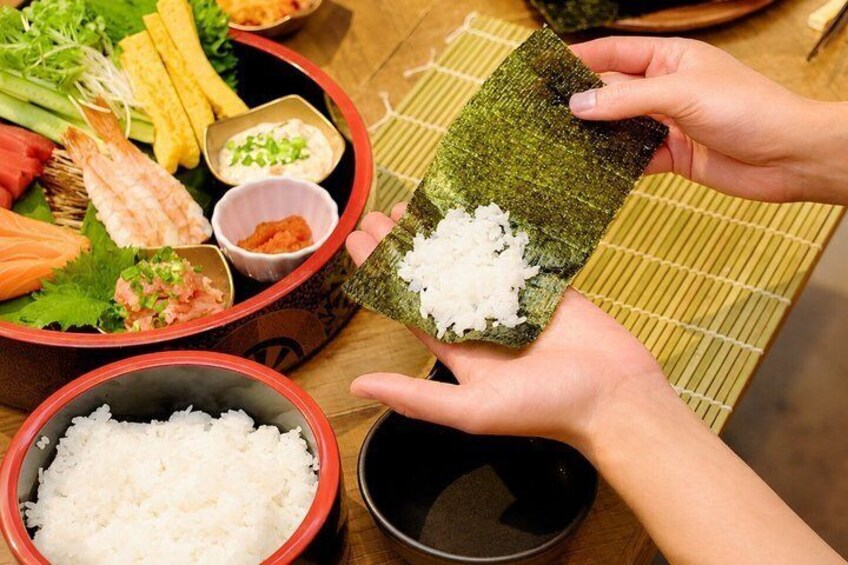 Une personne tenant un rouleau de sushi au bambou, garni de riz et d’algues, entourée de divers ingrédients pour sushi et de bols. 5