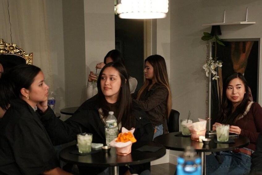 Un grupo de personas sentadas alrededor de mesas, con bebidas y comida, en una sala con candelabro y plantas decorativas. 4