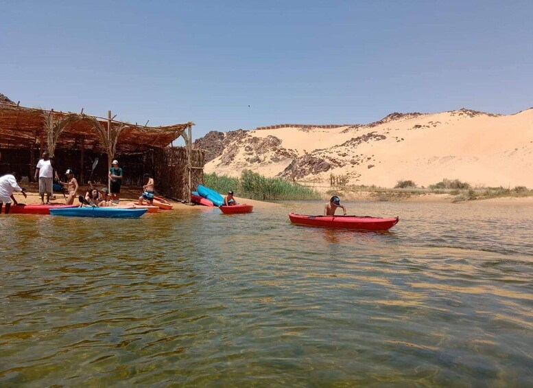 People in canoes on a calm river, a wooden structure on the bank, and sand dunes in the background. 4