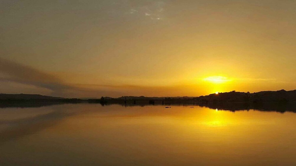 A calm lake at sunset with the sun reflecting on the water, surrounded by a hilly landscape. 3