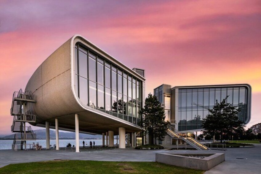 A modern building with a curved facade and a staircase leading to an upper level, situated by a waterfront. 1