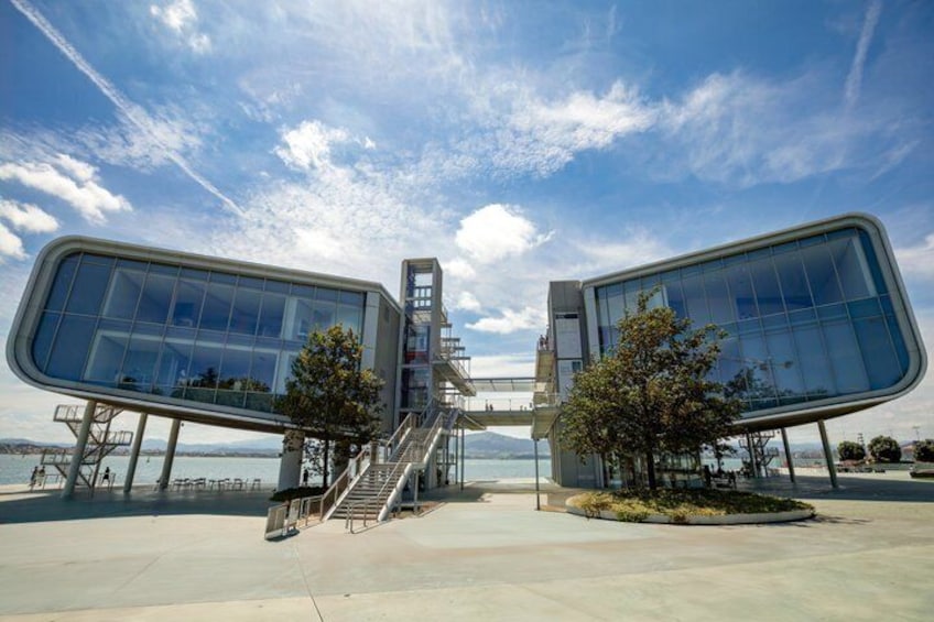 A modern architectural structure with glass panels and a staircase leading to an upper level, situated by a waterfront. 3