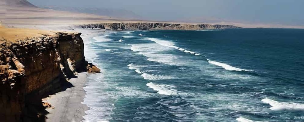 A coastal cliff with waves crashing against it, a sandy beach, and a distant mountain range. 5