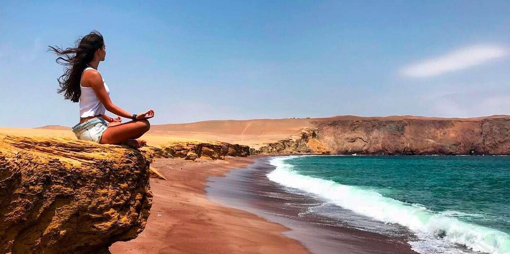A person meditating on a rock by a beach with clear turquoise water and sandy dunes. 4