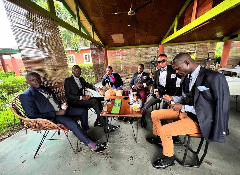 A group of men in formal attire seated around a table in an outdoor setting with a covered roof and greenery in the background. 2