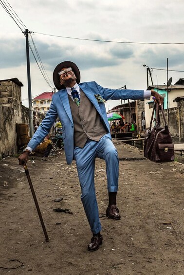 A person in a blue suit with a cane, holding a bag, in an outdoor setting with buildings and a cloudy sky. 3