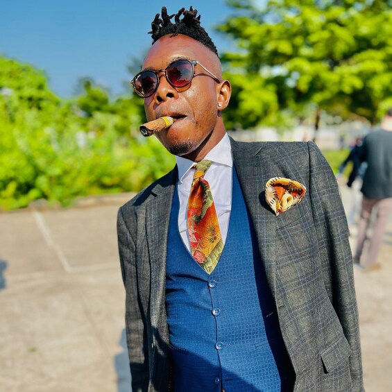 A man in a suit with a colorful tie and a pocket square, holding a cigar. 1