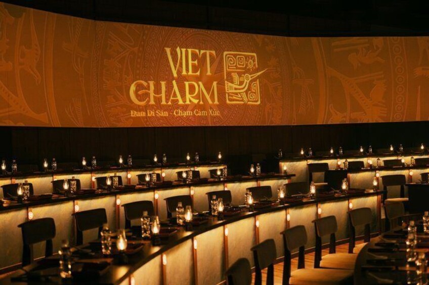 En svagt oplyst restaurant med dækkede borde, en væg med teksten 'VIET CHARM' og 'Bun Di San - Cham Cam Xuc', og en hyggelig atmosfære. 1