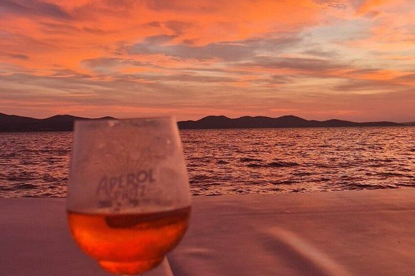 Et glas rødvin med 'Aperol' på, foran en solnedgang over vand med bjerge i baggrunden. 3