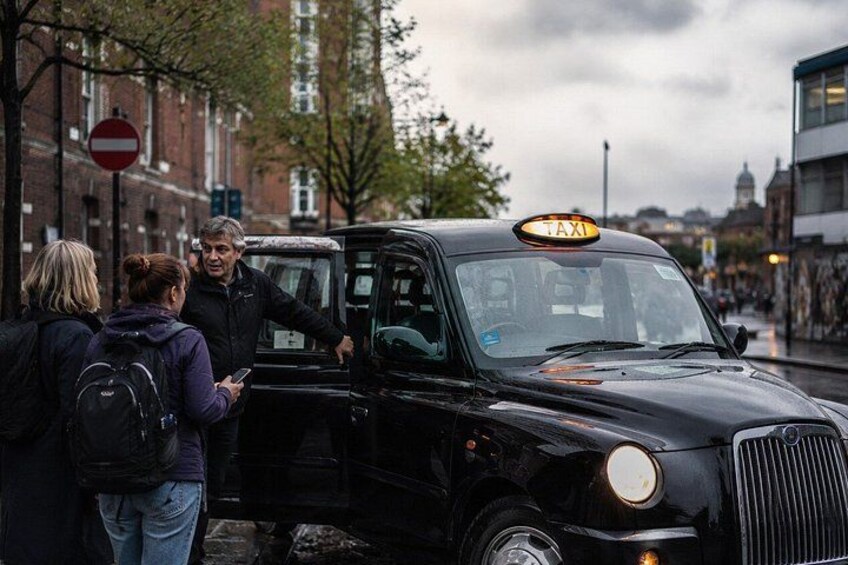 A black taxi with a 'TAXI' sign on top, parked on a city street with buildings and a 'no entry' sign in the background. 3