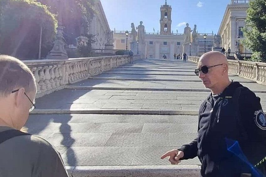 Un homme vêtu d’une veste noire, portant un insigne, se tient sur un pont de pierre. En arrière-plan, on aperçoit une fontaine et des statues. 1