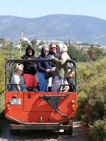 A group of people on a red vehicle with a license plate, wind turbines in the background, and a hilly landscape. 5