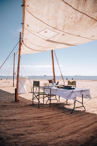A beach with a table set up under a sail, surrounded by chairs, and a wooden pole. 1