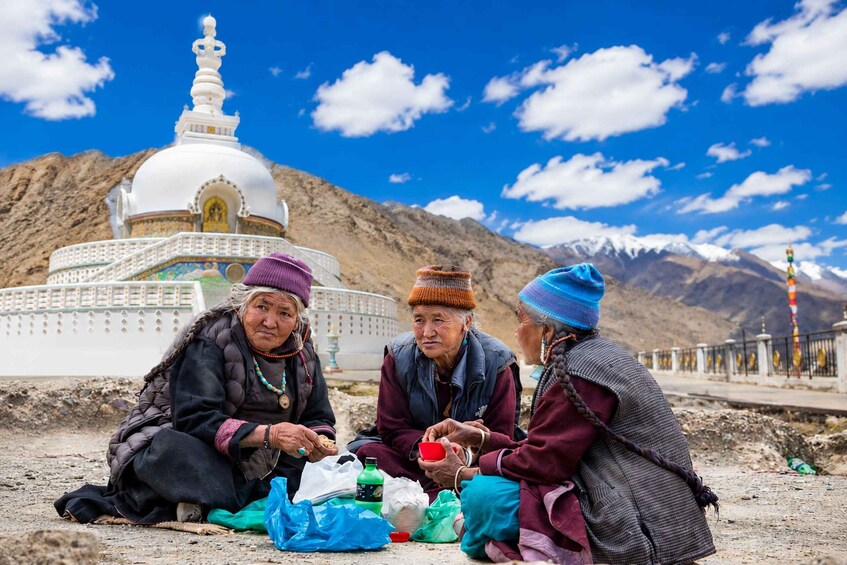 Three people sitting on the ground with a white stupa and mountains in the background. 1