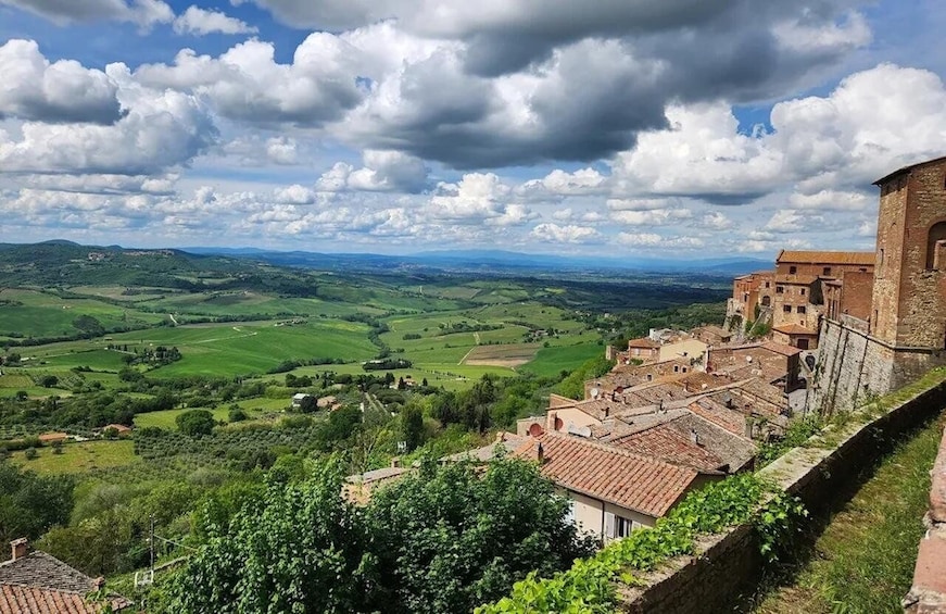 A historic town with terracotta-roofed buildings, a stone wall, and a panoramic view of rolling green hills under a partly cloudy sky. 1