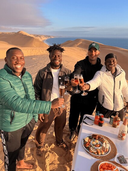 Four people toasting with drinks on a desert beach with a table of food and drinks. 2