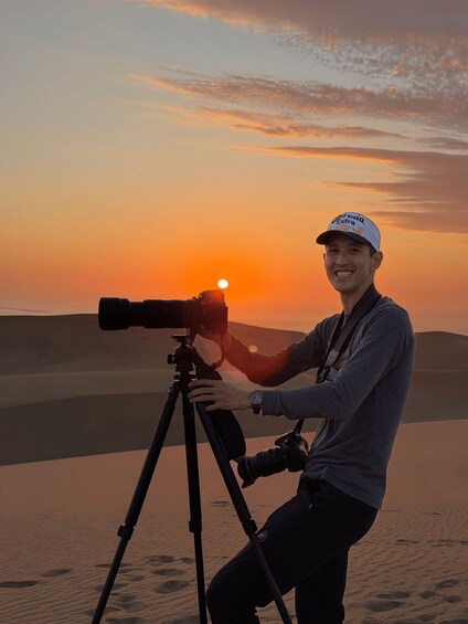 A person with a camera on a tripod, standing on sand dunes during sunset. 3
