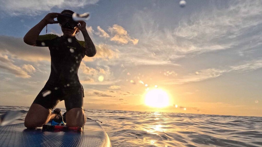 A person in a wetsuit kneeling on a surfboard with the sun setting in the background. 1