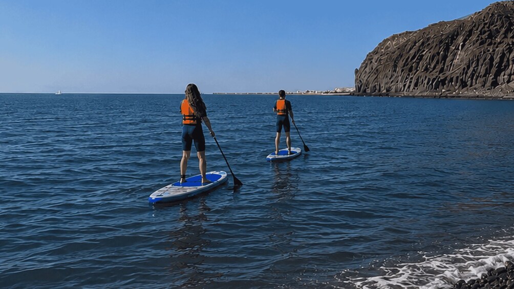 Two people paddleboarding on calm sea waters near a rocky coastline. 3