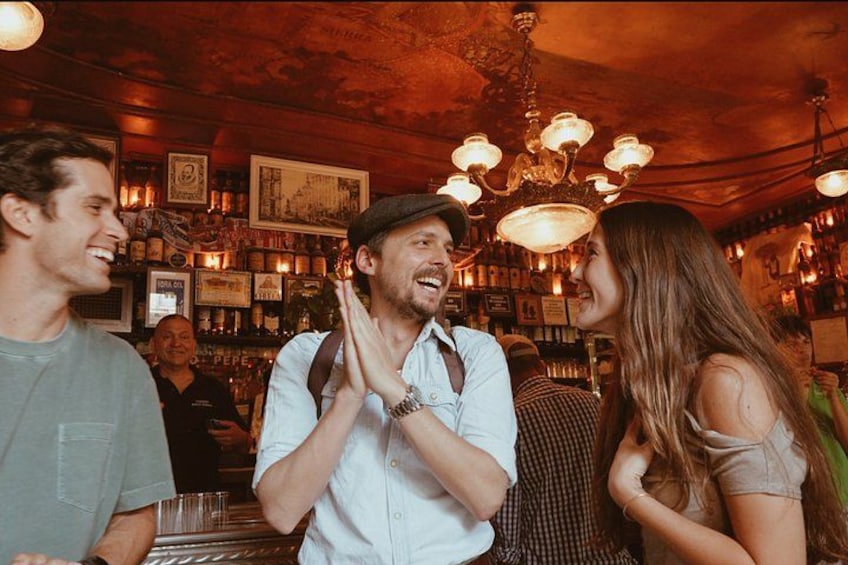 A bar with people socializing, a chandelier, and various framed pictures on the wall. 1
