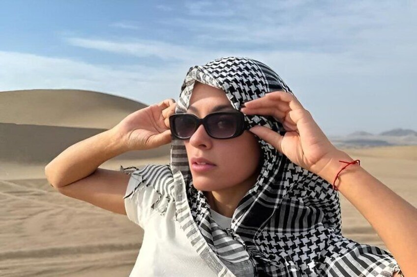 Een persoon met een geruit hoofddoek en zonnebril in een woestijnlandschap met zandduinen. 2
